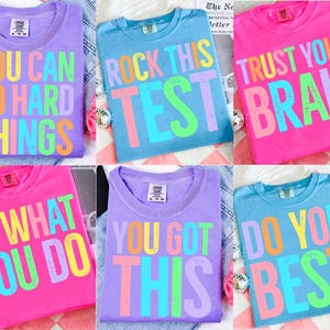 Può includere: Sei magliette colorate con frasi motivazionali. Le magliette sono in vari colori, tra cui viola, blu e rosa, con frasi come "You Can Do Hard Things" e "Trust Your Brain" stampate in lettere vivaci multicolori.