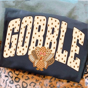 May include: A dark gray t-shirt featuring the word GOBBLE in large, cream-colored block letters with brown polka dots. Below the text is a graphic of a turkey with a patterned body, featuring brown and white gingham, leopard print, and floral details.
