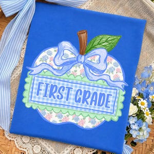 Op de afbeelding: Een felblauw shirt met een centrale appelvormige afbeelding met een bloemenpatroon, een lichtblauw gingham rechthoek met de tekst FIRST GRADE, een geschulpte lichtgroene rand, een bruine steel, een groen blad en een lichtblauwe strik.