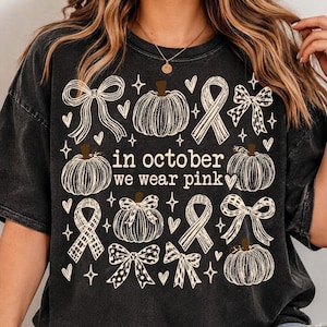 May include: A black mineral-washed t-shirt featuring a white graphic print of pumpkins, ribbons, and bows. The text in the center reads, "in october we wear pink." The design includes small heart and star accents scattered throughout the graphic.