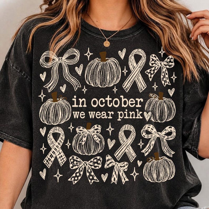May include: A black mineral-washed t-shirt featuring a white graphic print of pumpkins, ribbons, and bows. The text in the center reads, "in october we wear pink." The design includes small heart and star accents scattered throughout the graphic.