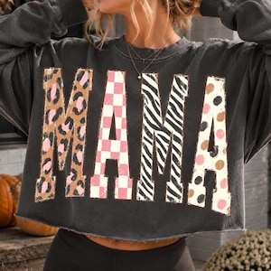 Peut inclure: Un sweat-shirt gris anthracite à manches longues avec un ourlet à bord brut. Le devant présente le mot MAMA en grandes lettres capitales, chacune avec un motif différent : léopard, damier rose et blanc, zébrures noires et blanches et pois.