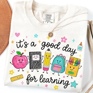 May include: A cream-colored t-shirt featuring the text "it's a good day for learning" in black script. The graphic includes cartoon school supplies with faces: a pink apple, a black notebook, a yellow pencil, a teal backpack, and a crayon box.