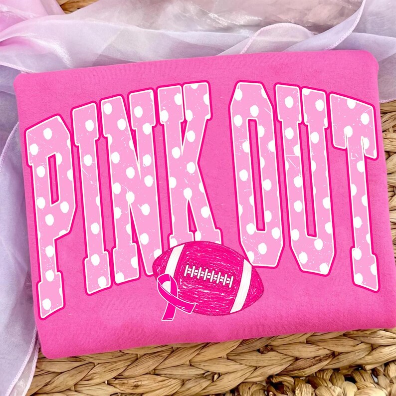 May include: A bright pink sweatshirt featuring the text PINK OUT in large, white polka-dotted block letters. Below the text is a graphic of a pink football with white laces and a pink awareness ribbon attached to the side of the football.
