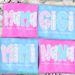 Puede incluir: Cuatro camisetas coloridas con los nombres "MOM", "GIGI", "MIMI" y "NINI" en un diseño floral. Cada camiseta tiene un nombre debajo, como "Layla Oliver Taylor". Las camisetas son rosas y azules.