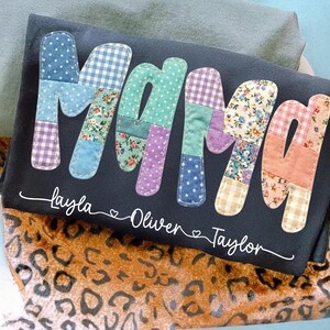 May include: Black pillow with the word "Mama" in patchwork letters. The letters are made of various patterned fabrics in blue, green, purple, and pink. Below the word are the names Layla, Oliver, and Taylor.
