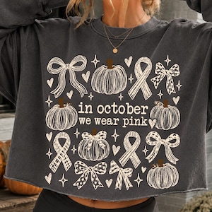 May include: A charcoal gray long-sleeve sweatshirt featuring a cream-colored graphic print of pumpkins, bows, and awareness ribbons. The text "in october we wear pink" is centered among the illustrations, surrounded by small heart and star icons.