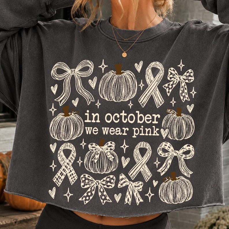 May include: A charcoal gray long-sleeve sweatshirt featuring a cream-colored graphic print of pumpkins, bows, and awareness ribbons. The text "in october we wear pink" is centered among the illustrations, surrounded by small heart and star icons.