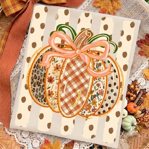 May include: A rectangular sign featuring a central pumpkin illustration with a light pink bow. The pumpkin segments display leopard print, orange floral patterns, and orange gingham. The background has beige vertical stripes with brown polka dots.