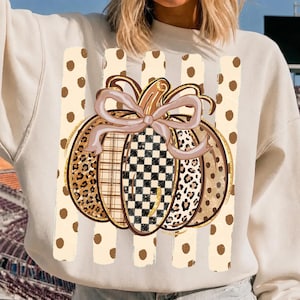 K&ouml;nnte beinhalten: Ein cremefarbenes Sweatshirt mit einer zentralen Grafik eines K&uuml;rbisses mit Schleife. Der K&uuml;rbis hat Segmente mit Leopardenmuster, Karomuster und Schachbrettmuster. Der Hintergrund der Grafik besteht aus vertikalen Streifen mit braunen Punkten.