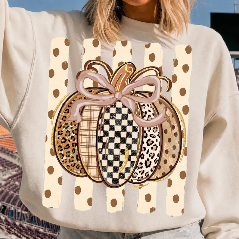 K&ouml;nnte beinhalten: Ein cremefarbenes Sweatshirt mit einer zentralen Grafik eines K&uuml;rbisses mit Schleife. Der K&uuml;rbis hat Segmente mit Leopardenmuster, Karomuster und Schachbrettmuster. Der Hintergrund der Grafik besteht aus vertikalen Streifen mit braunen Punkten.