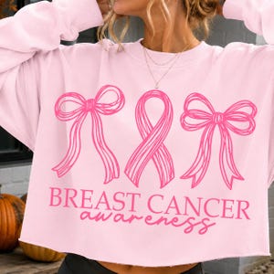 May include: A light pink long-sleeve sweatshirt featuring a dark pink graphic print of two bows flanking a central awareness ribbon. Below the graphic, the text reads BREAST CANCER awareness in a combination of block and script lettering.