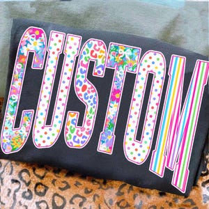 May include: A black t-shirt with the word "CUSTOM" in a large, colorful, outlined font. The letters are decorated with floral, polka dot, and striped patterns. The shirt is folded on a leopard print surface.