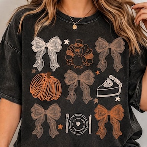 Puede incluir: Una camiseta negra con efecto lavado al ácido que presenta un estampado gráfico de cuatro lazos, un pavo, una calabaza, un trozo de tarta y un cubierto con tenedor y cuchillo. El diseño incluye pequeñas estrellas en tonos naranja, marrón y blanco.