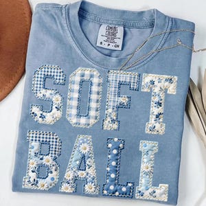 May include: A light blue Comfort Colors t-shirt with the word "SOFTBALL" appliqued in various blue and white floral and gingham patterns. The shirt has a crew neck and short sleeves. A small tag is visible near the neckline.