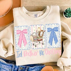 Puede incluir: Sudadera color crema con un dise&ntilde;o gr&aacute;fico. Presenta un lazo rosa, un carrito de compras lleno de art&iacute;culos vintage y un lazo azul. El texto "I'd Rather Be Thrifting" est&aacute; impreso debajo.