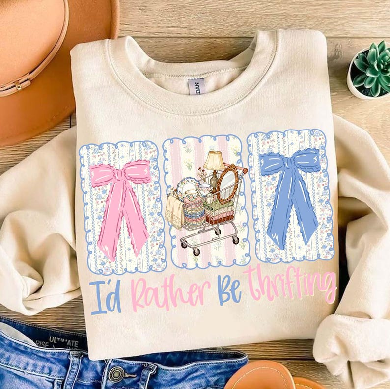 Puede incluir: Sudadera color crema con un dise&ntilde;o gr&aacute;fico. Presenta un lazo rosa, un carrito de compras lleno de art&iacute;culos vintage y un lazo azul. El texto "I'd Rather Be Thrifting" est&aacute; impreso debajo.