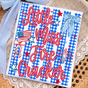 May include: White square fabric with a blue and white gingham pattern. The text "Little Miss Fire Cracker" is written in red script. The design includes a bow, stars, a firework, and an American flag.
