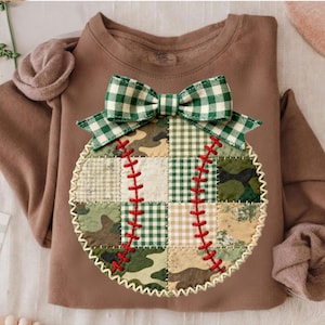 Puede incluir: Una sudadera marrón con un aplique de béisbol. El diseño de béisbol está hecho de tela de retazos en patrones de camuflaje, cuadros verdes y beige. Un lazo de cuadros verdes y blancos se encuentra encima del béisbol, con costuras rojas.