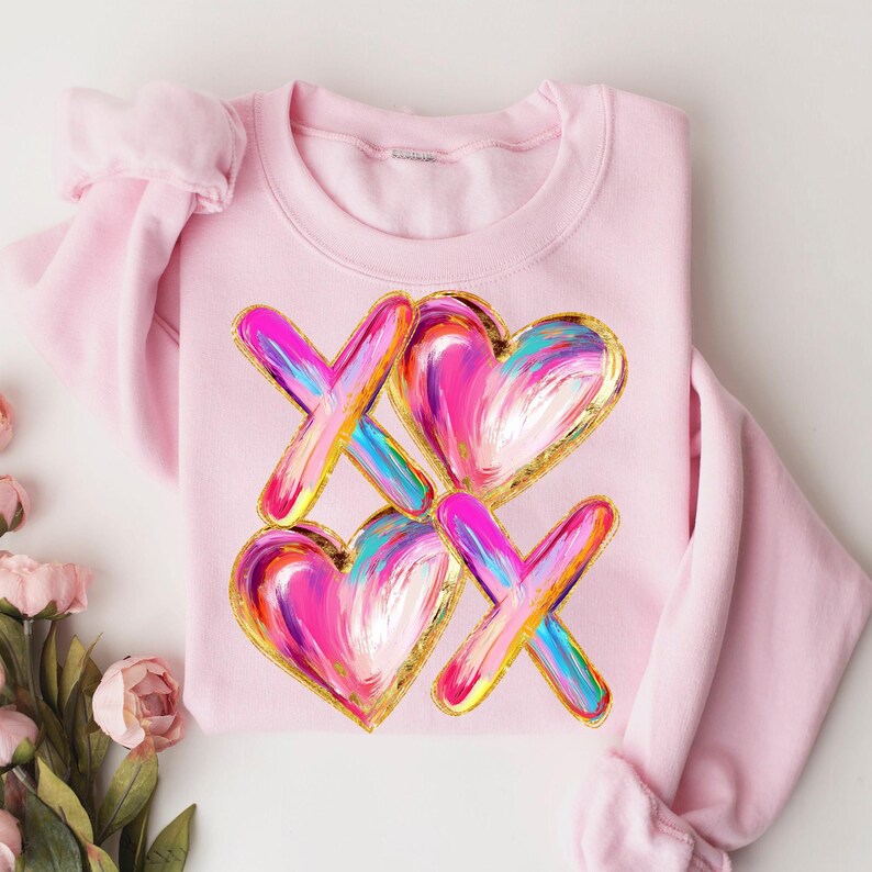 Puede incluir: Sudadera rosa con un dise&ntilde;o gr&aacute;fico de corazones y X coloridos con detalles dorados. El dise&ntilde;o recuerda a una pintura.