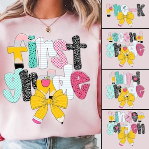 Könnte beinhalten: Rosafarbener Pullover mit einem grafischen Design der Wörter "first grade" in bunten, gemusterten Buchstaben mit einer gelben Schleife und einem Bleistift. Das Design beinhaltet einen roten Apfel, karierte Muster und Punkte.
