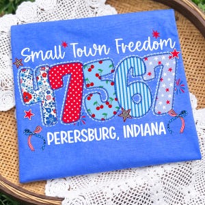 May include: Blue t-shirt with the text "Small Town Freedom" and the numbers "47361" in a patchwork design. The numbers are decorated with floral, polka dot, cherry, and star patterns. The text "PERERSBURG, INDIANA" is below the numbers.