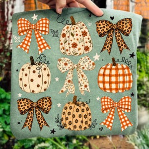 May include: A sage green t-shirt featuring a repeating pattern of pumpkins and bows. The pumpkins include floral, polka dot, and plaid designs. The bows feature orange gingham, leopard print, and floral patterns, accented by small white stars.