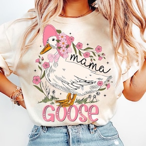 Pu&ograve; includere: Una maglietta bianca con una grafica di un'oca che indossa un cappello rosa e fiori. Il testo "mama goose" &egrave; stampato sulla maglietta.
