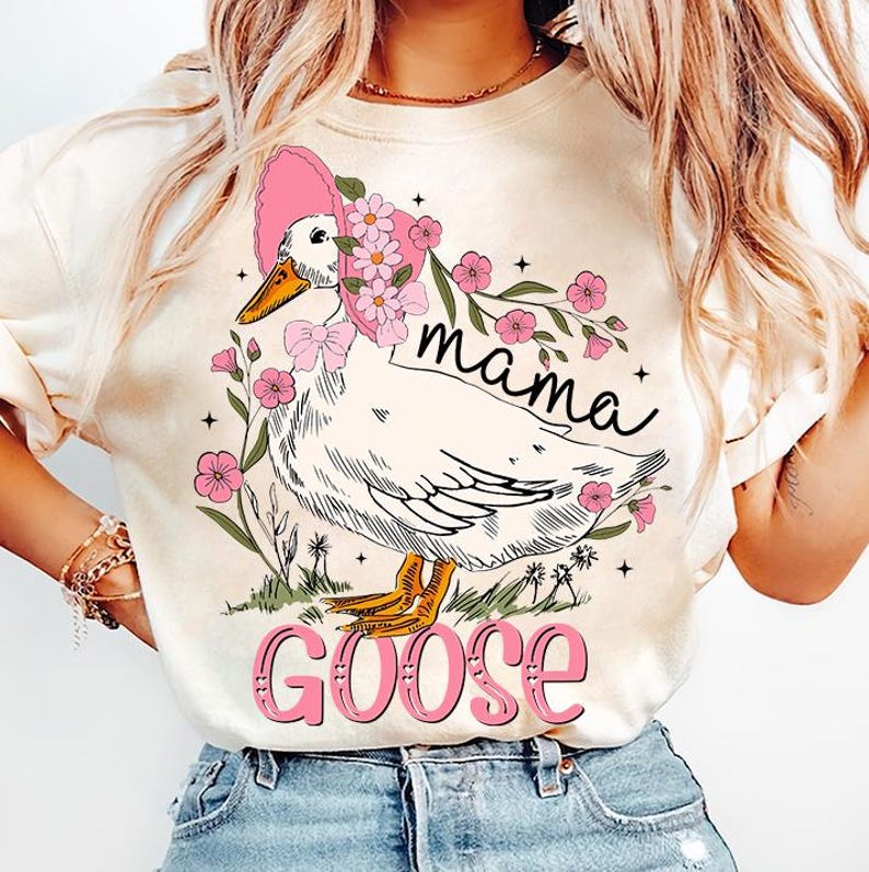 Pu&ograve; includere: Una maglietta bianca con una grafica di un'oca che indossa un cappello rosa e fiori. Il testo "mama goose" &egrave; stampato sulla maglietta.