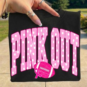 May include: A black t-shirt featuring the text PINK OUT in large, pink, polka-dotted block letters. Below the text is a pink American football graphic with a pink awareness ribbon integrated into the design, all printed on the front of the shirt.