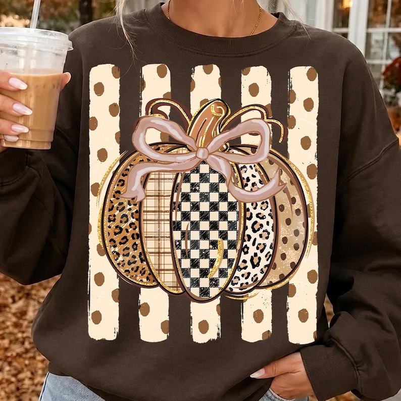 K&ouml;nnte beinhalten: Ein dunkelbraunes Sweatshirt mit Rundhalsausschnitt und einem zentralen K&uuml;rbis-Motiv mit einer hellrosa Schleife. Der K&uuml;rbis besteht aus Segmenten mit Leopardenmuster, Karomuster und schwarz-wei&szlig;em Schachbrettmuster auf beigen Streifen.