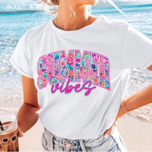 K&ouml;nnte beinhalten: Wei&szlig;es T-Shirt mit einem floralen Druckdesign, auf dem "SUMMER vibes" in rosa Buchstaben steht.