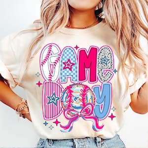 May include: A white t-shirt with a pink, blue, and white graphic design that says "GAME DAY" with a baseball and a bow.