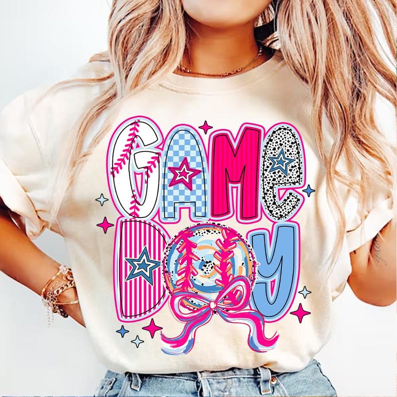 May include: A white t-shirt with a pink, blue, and white graphic design that says "GAME DAY" with a baseball and a bow.