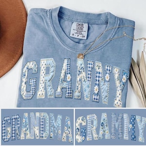 May include: A light blue Comfort Colors t-shirt with the word "GRANNY" appliqued across the front in a patchwork design of blue floral and gingham fabrics. A gold necklace with a pendant rests on the shirt. The shirt is folded.