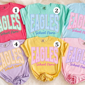 Pode incluir: Seis camisetas de manga curta e gola redonda em várias cores pastel, cada uma com a palavra EAGLES em uma fonte universitária grande com o texto School Name abaixo, acentuadas por pequenos gráficos de estrelas e numeradas de um a seis.