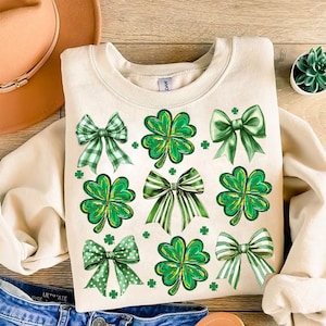 May include: A cream-coloured sweatshirt with a green shamrock and bow design. The design features multiple shamrocks and bows in a repeating pattern.