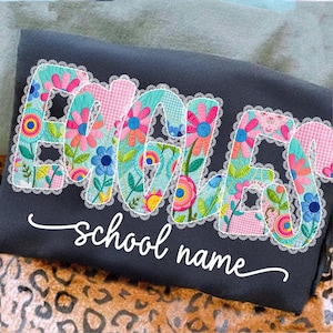 May include: A dark gray sweatshirt with the name "EMMA" embroidered in colorful floral patterns and outlined with white lace. The words "school name" are written in cursive below the name.