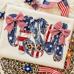 May include: Cream-coloured t-shirt featuring the word "USA" appliquéd in denim, American flag fabric, and leopard print. Each letter is decorated with a red and white striped bow. Patriotic design.