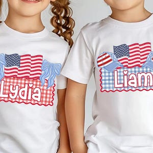Puede incluir: Camisetas blancas con dise&ntilde;os patri&oacute;ticos. Una camiseta presenta el nombre "Lydia" con lazos azules y una bandera americana. La otra camiseta tiene el nombre "Liam" con gorras de b&eacute;isbol y una bandera americana.