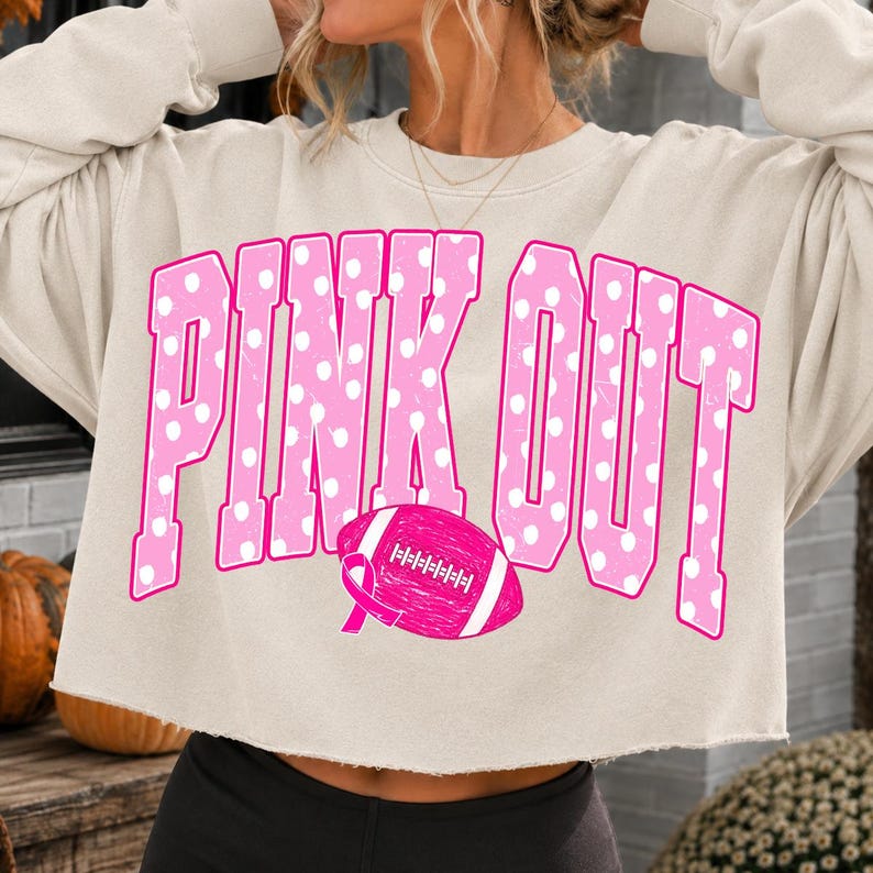 May include: A beige cropped sweatshirt featuring the text PINK OUT in large pink letters with white polka dots. Below the text is a pink football graphic with a breast cancer awareness ribbon integrated into the design on the left side.