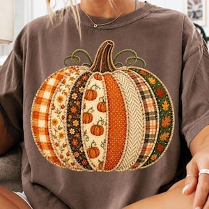 Puede incluir: Una camiseta de color marrón oscuro con un gráfico central grande de una calabaza compuesta por varios patrones de tela verticales, incluyendo cuadros naranjas, estampados florales, lunares, espiga y motivos de hojas verdes con un tallo marrón.