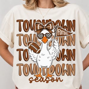 May include: A cream-colored t-shirt featuring a graphic of a white duck wearing a brown bow and checkered heart-shaped sunglasses. The duck holds a football and a pennant labeled FOOTBALL. The background text reads TOUCHDOWN SEASON.