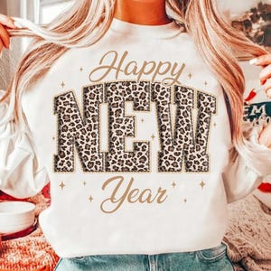 K&ouml;nnte beinhalten: Wei&szlig;es Sweatshirt mit dem Schriftzug "Happy NEW Year" in goldener Schrift und Leopardenmuster-Blockbuchstaben. Das Design enth&auml;lt goldene Sternakzente. Ein festliches Oberteil zum Feiern des neuen Jahres.