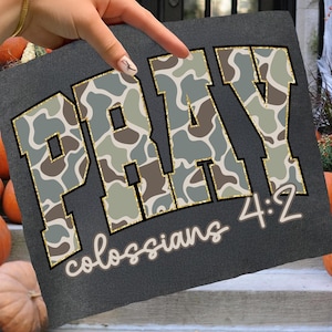 May include: Dark gray t-shirt with the word "PRAY" in a camouflage pattern with gold trim. The text "Colossians 4:2" is written below in a cursive font. The shirt is displayed on a set of steps with pumpkins in the background.