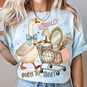 May include: Light blue t-shirt with a goose illustration pushing a shopping cart filled with vintage items. The text "LITERALLY BORN TO THRIFT" is printed above and below the goose. The goose wears a blue bow.