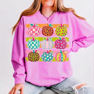 May include: A bright pink long-sleeve sweatshirt featuring a graphic print of nine pumpkins arranged in a grid. Each pumpkin displays a unique pattern, including checkered, leopard print, and polka dot designs in vibrant neon colors like green and orange.