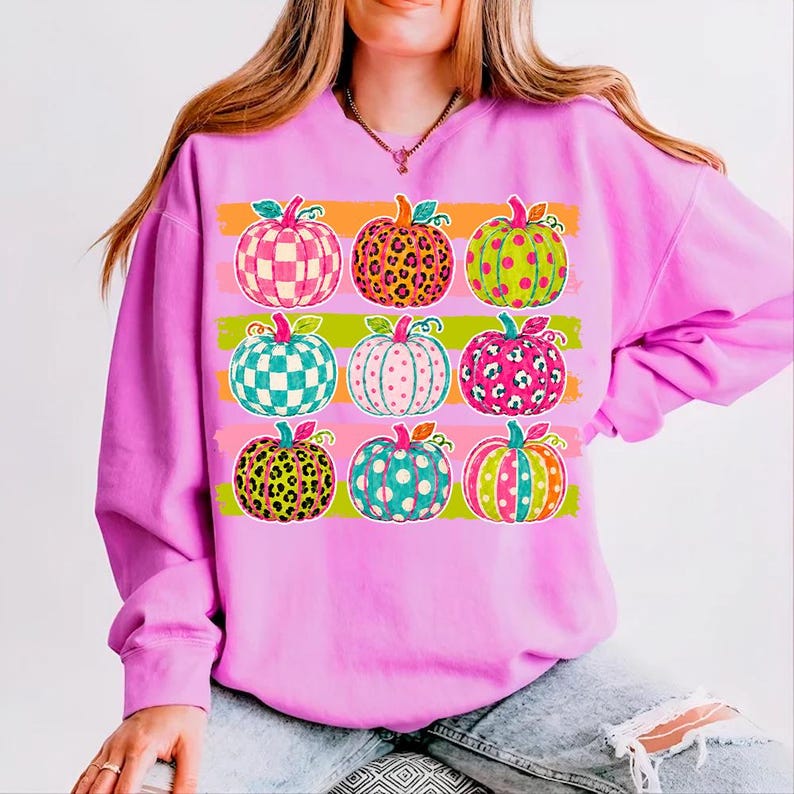 May include: A bright pink long-sleeve sweatshirt featuring a graphic print of nine pumpkins arranged in a grid. Each pumpkin displays a unique pattern, including checkered, leopard print, and polka dot designs in vibrant neon colors like green and orange.
