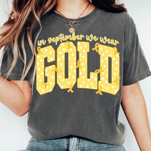 May include: A charcoal gray short-sleeve t-shirt featuring the yellow text In September We Wear Gold. The word GOLD is printed in large block letters with a white polka dot pattern and small yellow awareness ribbons scattered throughout.
