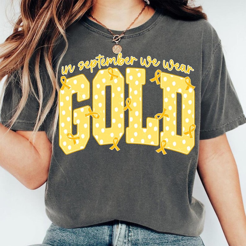 May include: A charcoal gray short-sleeve t-shirt featuring the yellow text In September We Wear Gold. The word GOLD is printed in large block letters with a white polka dot pattern and small yellow awareness ribbons scattered throughout.